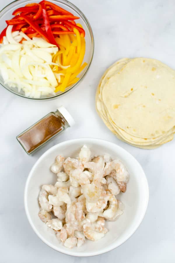 a bowl of uncooked shrimp, a bowl of sliced peppers and onions, and a stack of tortillas