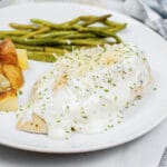 closeup of herb chicken covered with creamy sauce on a plate with potatoes and green beans