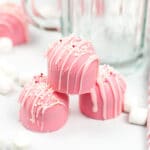 2 pink valentines day hot cocoa bombs stacked in front of a glass mug