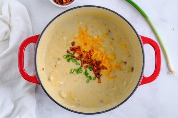 a dutch oven pot with potato soup topped with bacon, shredded cheese and scallions