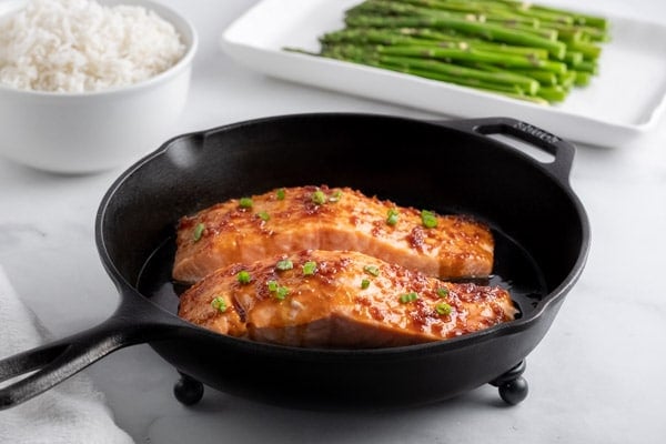 two salmon fillets in a cast iron pan with a bowl of white rice and a plate of asparagus in the background