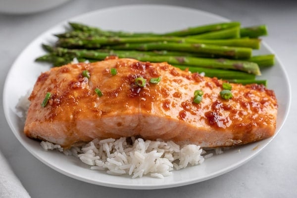 a white plate with a salmon fillet over white rice and a side of asparagus