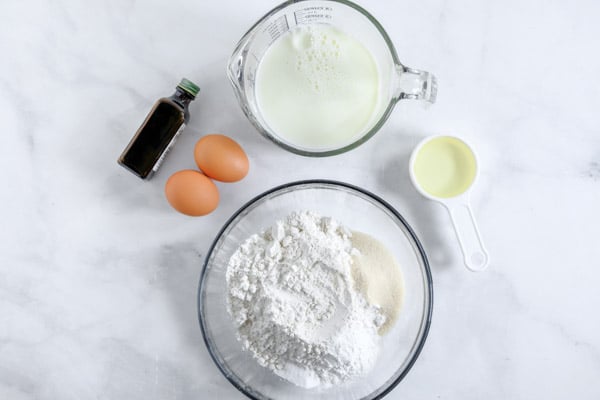 a bowl with flour, a bowl of milk a measuring cup with oil, two eggs and bottle of vanilla