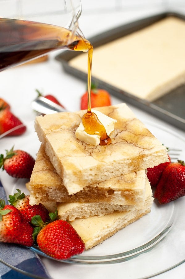 syrup being poured over a stack of sheet pan pancakes with strawberries on the plate