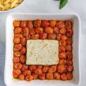 square white baking dish with feta cheese block, italian seasoning and cooked cherry tomatoes