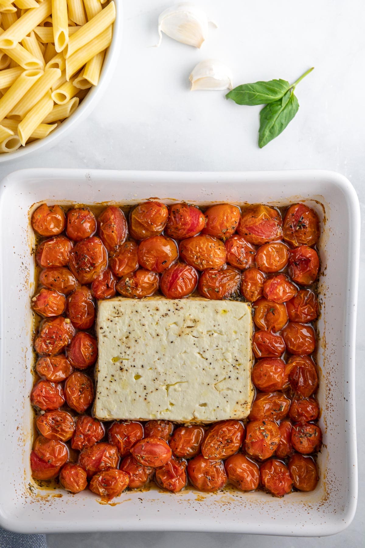 a white baking dish with cooked cherry tomatoes with a block of feta cheese in the center