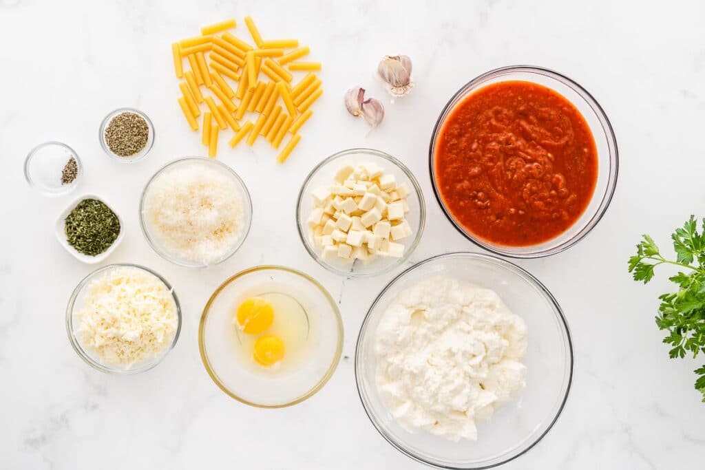 several glass bowls with ingredients for baked ziti tomato sauce, cheeses, pasta, eggs and spices