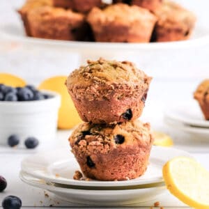 two blueberry lemon muffins on a white plate with a cake stand in the background and blueberries and lemon slices on the counter