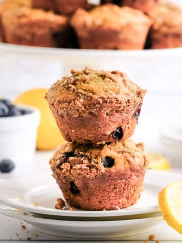 two blueberry lemon muffins on a white plate with a cake stand in the background and blueberries and lemon slices on the counter