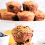 a white plate with two blueberry muffins stacked and a white cake stand in the background with five muffins