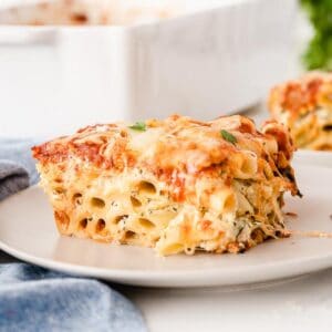 slice of cheesy baked ziti on a white plate with a blue linen napkin beside