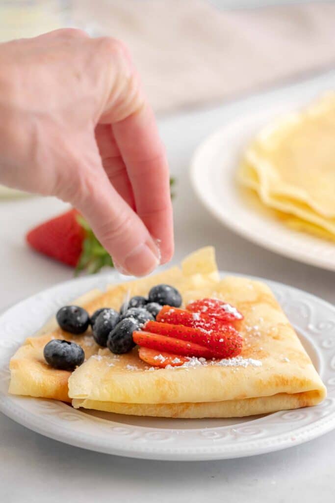 a womans hand sprinkling powdered sugar over crepes