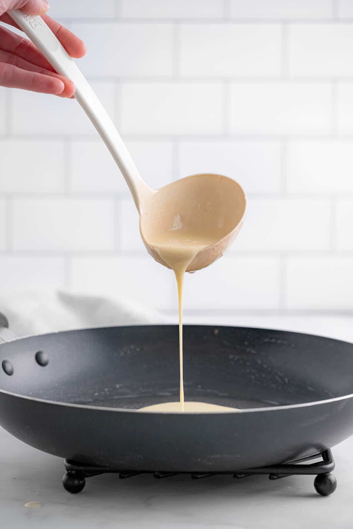 crepe batter being poured out of a white ladle into a black pan