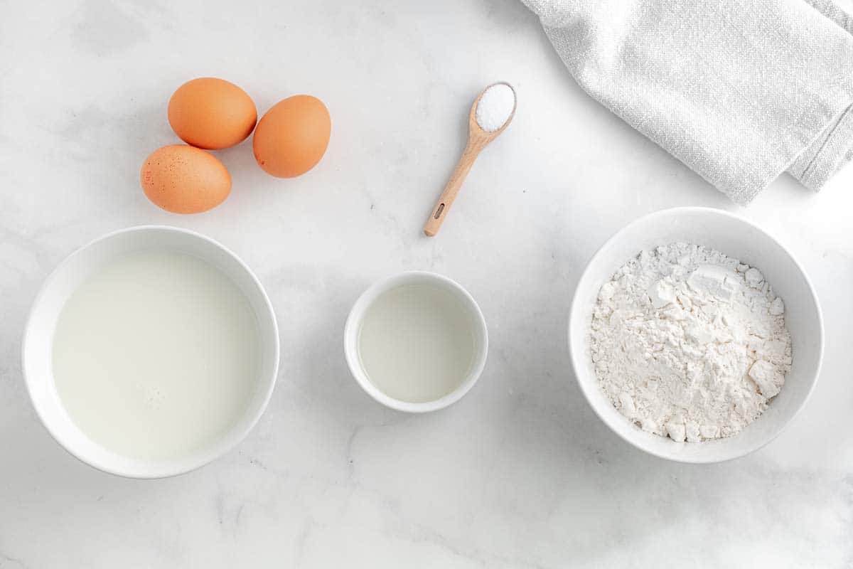 three white bowls filled with ingredients for homemade crepes, three eggs and a wooden measuring spoon