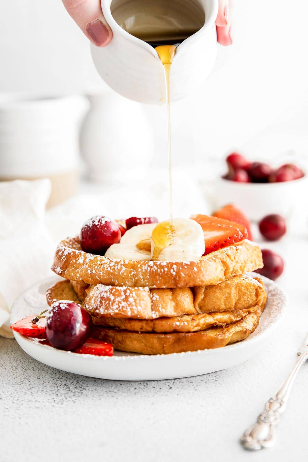 syrup being poured over a stack of french toast topped with slices of banana, strawberries and cherries