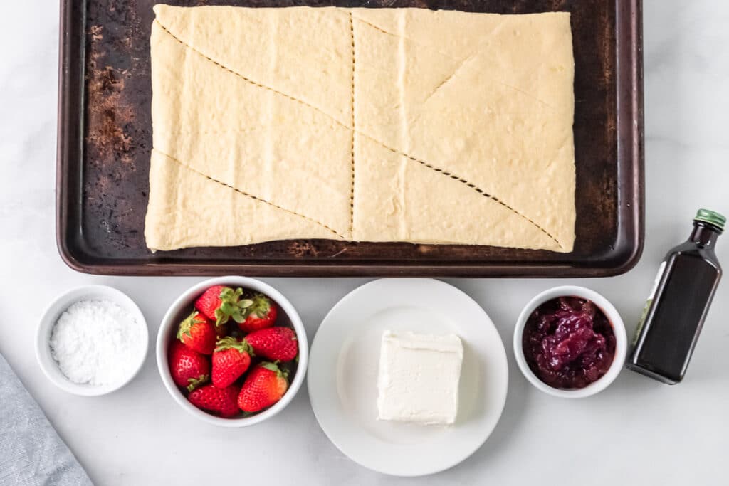 uncooked crescent roll dough triangles on a baking sheet, strawberries, strawberry jam, powdered sugar in white bowls, and cream cheese on plates