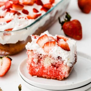 closeup of a square slice of strawberry poke cake topped with fresh strawberries on a white plate
