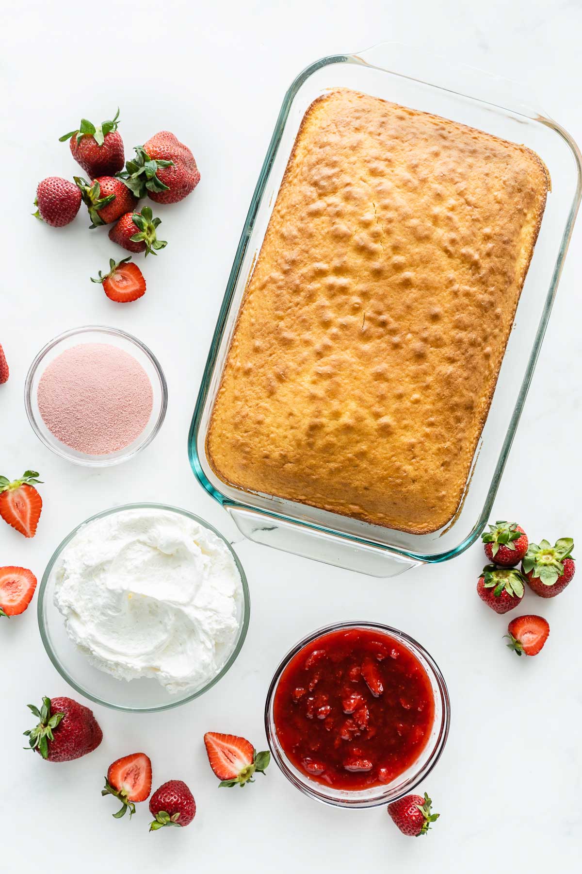 rectangle glass baking dish with white cake and three glass bowls with strawberries, whipped cream and jello mix powder