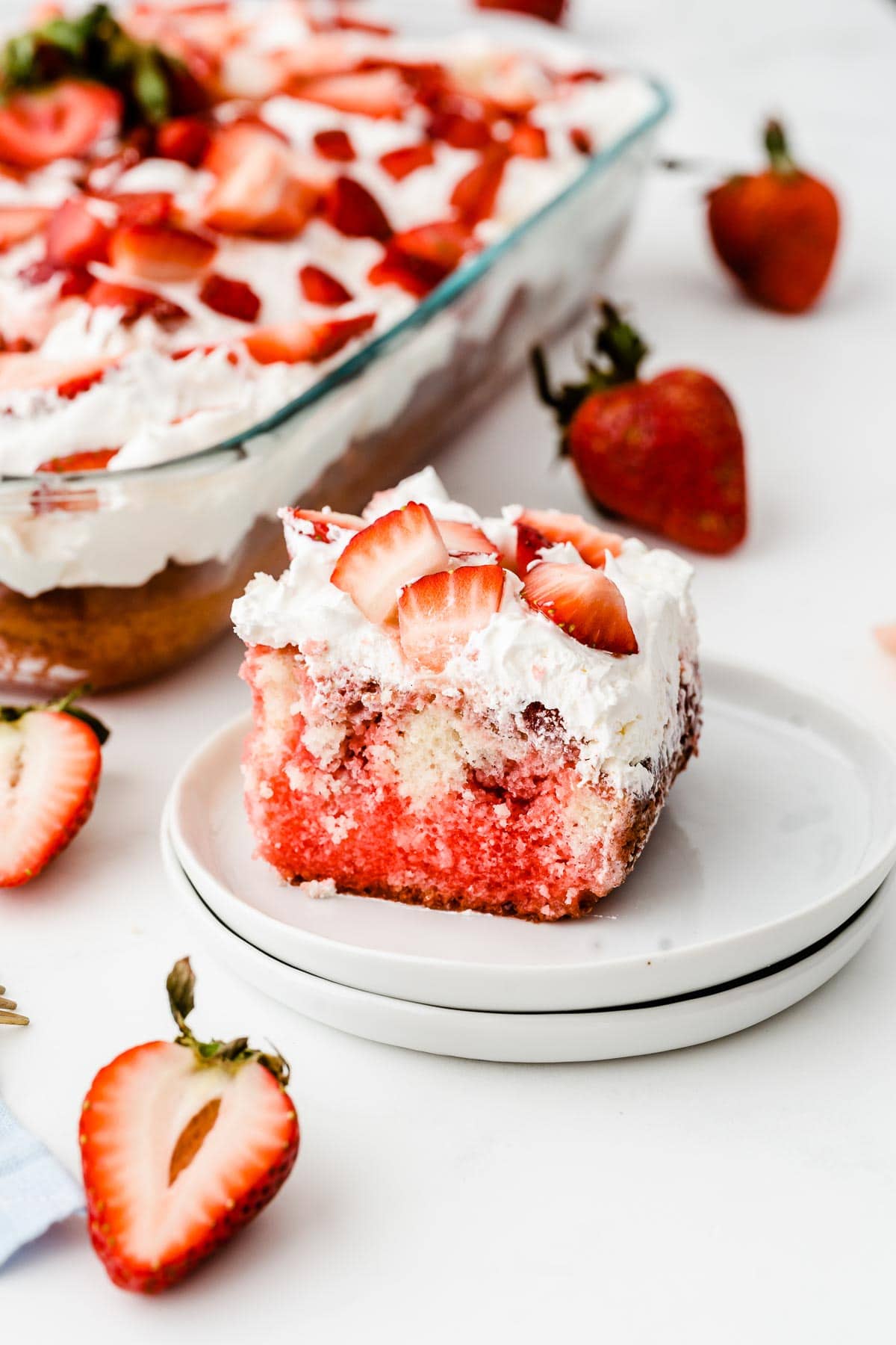a white plate with a slice of strawberry poke cake with a whole cake dish in the background