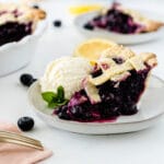 a slice of blueberry pie with lattice top with a scoop of vanilla ice cream on a white plate