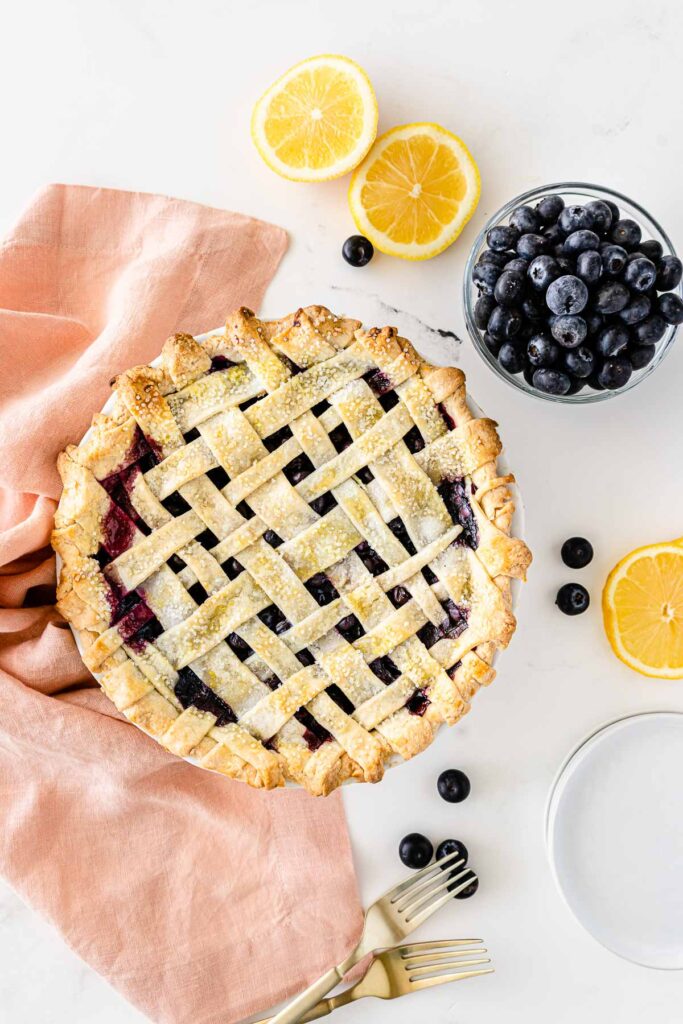 whole homemade blueberry pie with a lattice top layer of crust surrounded by white plates, blueberries and lemon slices