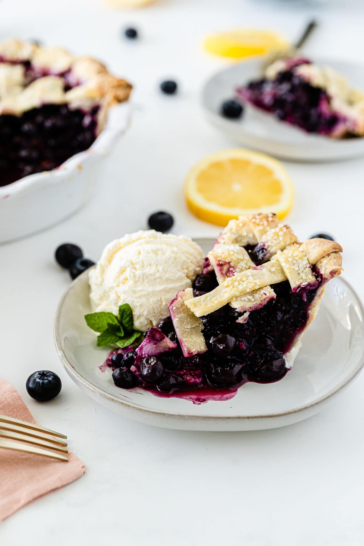 a white plate with a slice of blueberry pie with a scoop of ice cream surrounded by lemons and fresh blueberries