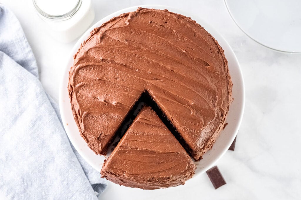 a chocolate frosted cake on a white cake stand with a triangle piece cut out but still on the stand