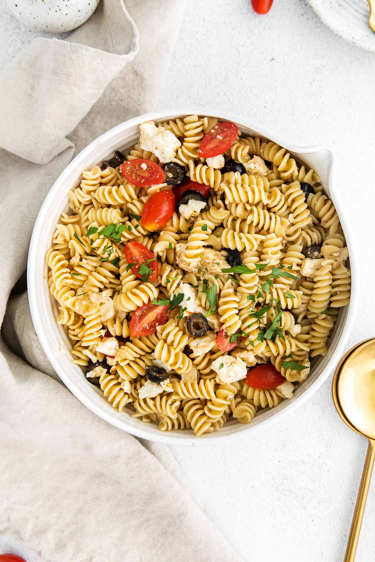 white bowl with greek pasta salad mixed with cherry tomatoes, olives and feta cheese with a tan napkin and gold serving spoon