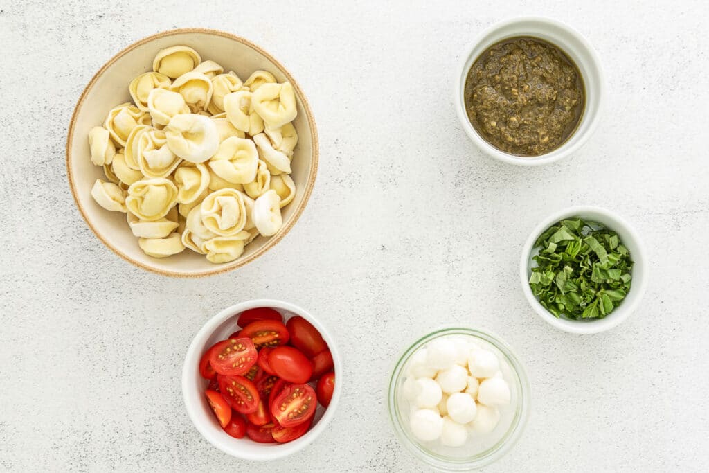 small bowls of tortellini pasta, pesto, cherry tomatoes, mozzarella cheese balls, and fresh basil