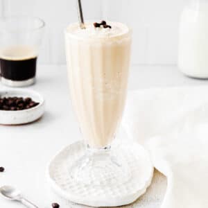 coffee milkshake in tall glass with a straw with a cup of coffee and milk in the background