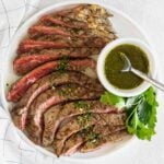 sliced flank steak with a small bowl of chimichurri sauce on the side on a white plate