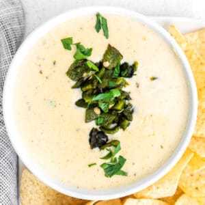 closeup of queso dip with diced poblano peppers in a white bowl surrounded by tortilla chips