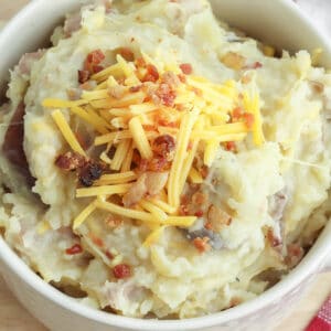 closeup of a white bowl of mashed potatoes loaded with bacon, and shredded cheese.