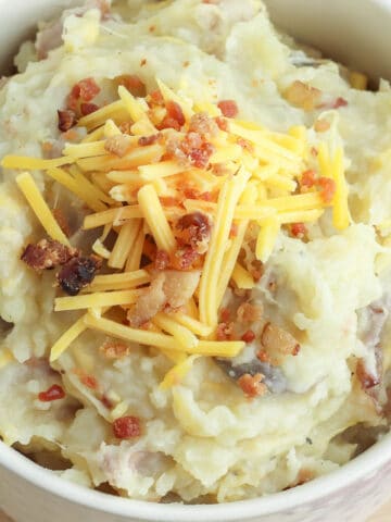 closeup of a white bowl of mashed potatoes loaded with bacon, and shredded cheese.