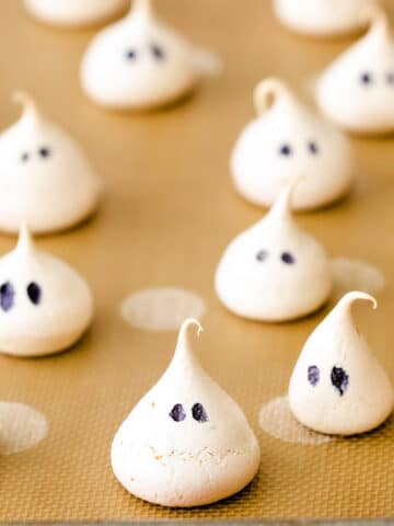 several little ghost shaped meringue cookies on a brown baking mat