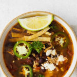 white bowl of soup with tomato topped with tortilla strips, jalapeños, cheese in a