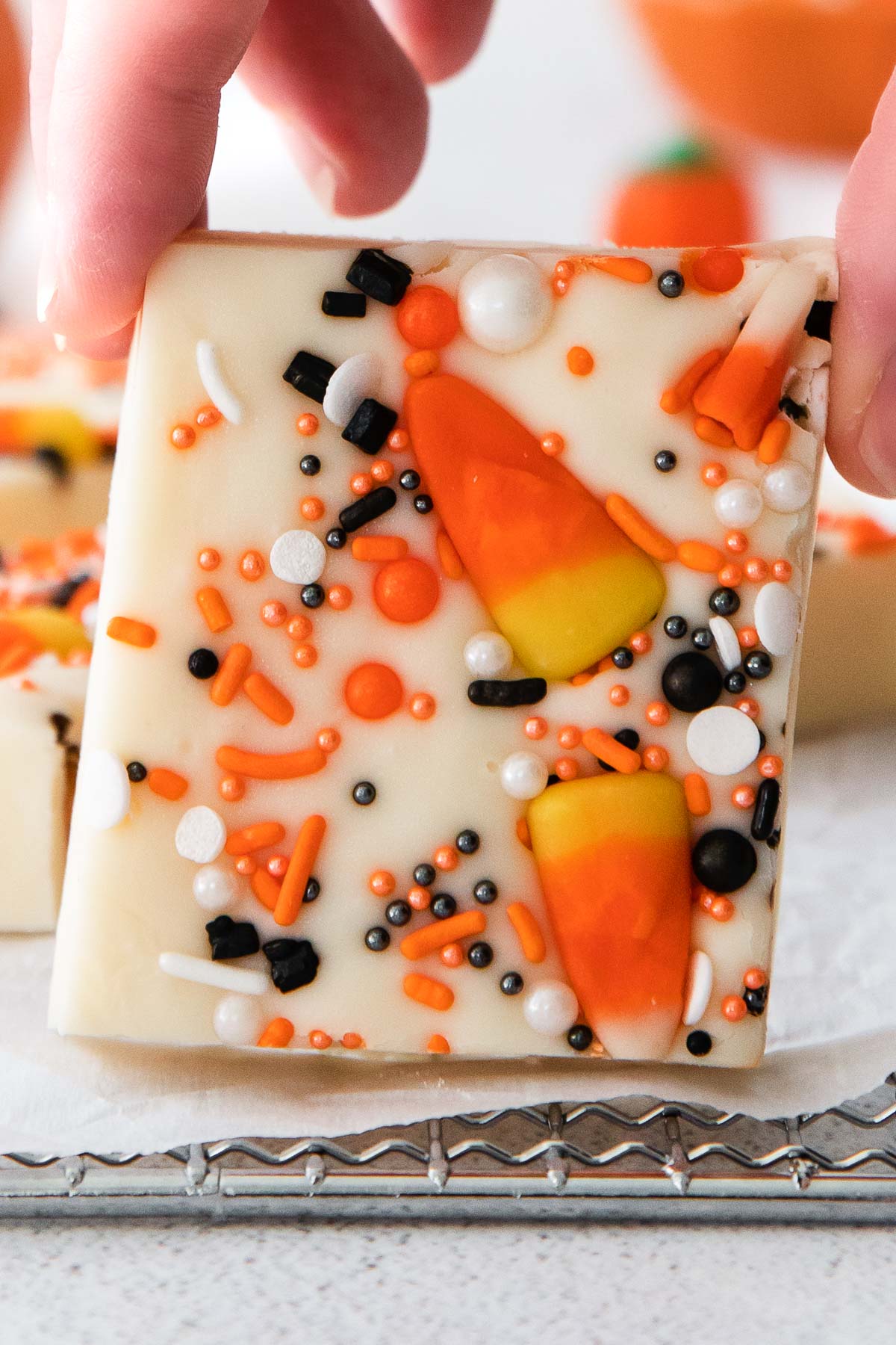 closeup of a square of white fudge topped with white, orange and black round sprinkles and candy corn being held by fingers