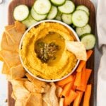 bowl of hummus with pumpkin seeds and olive oil on a dark wood cutting board with pita chips, cucumbers and carrots