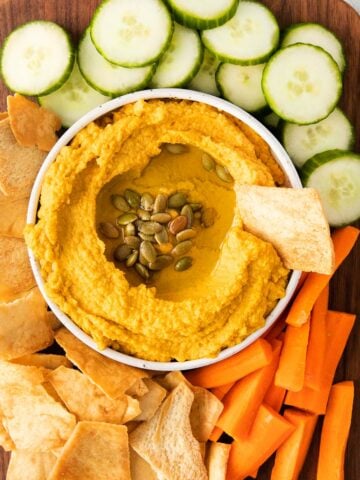 bowl of hummus with pumpkin seeds and olive oil on a dark wood cutting board with pita chips, cucumbers and carrots