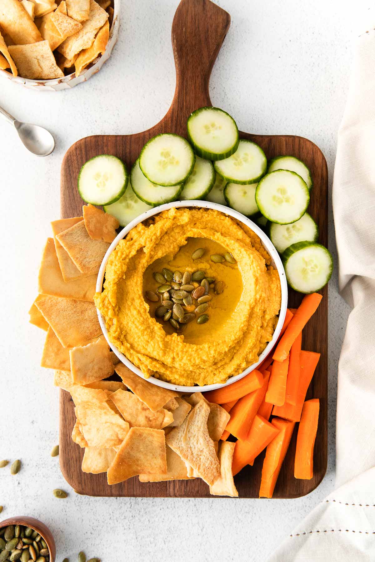 white bowl of hummus topped with pumpkin seeds on a wood cutting board surrounded by fresh carrots, sliced and bagel chips