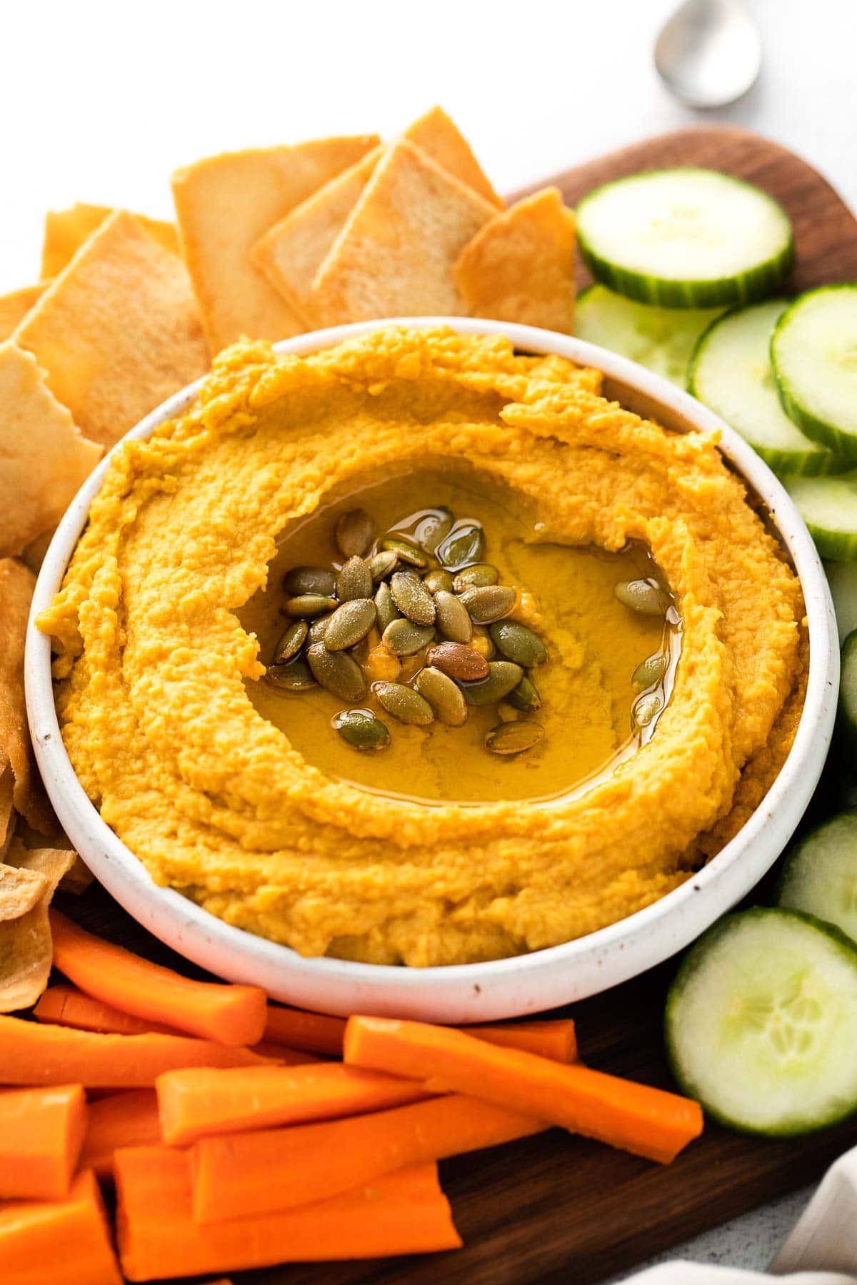 closeup of a white bowl of pumpkin hummus topped with pumpkin seeds surrounded by sliced carrots and cucumbers