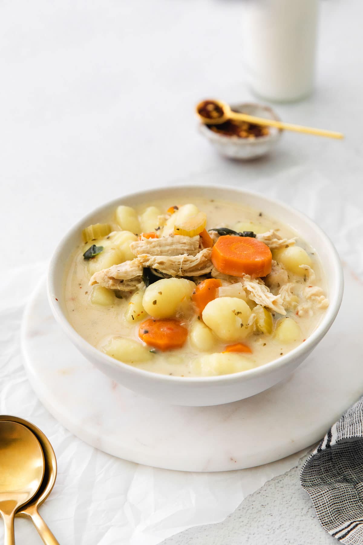 white bowl of chicken and gnocchi soup with gold spoons and a black and white checked napkin