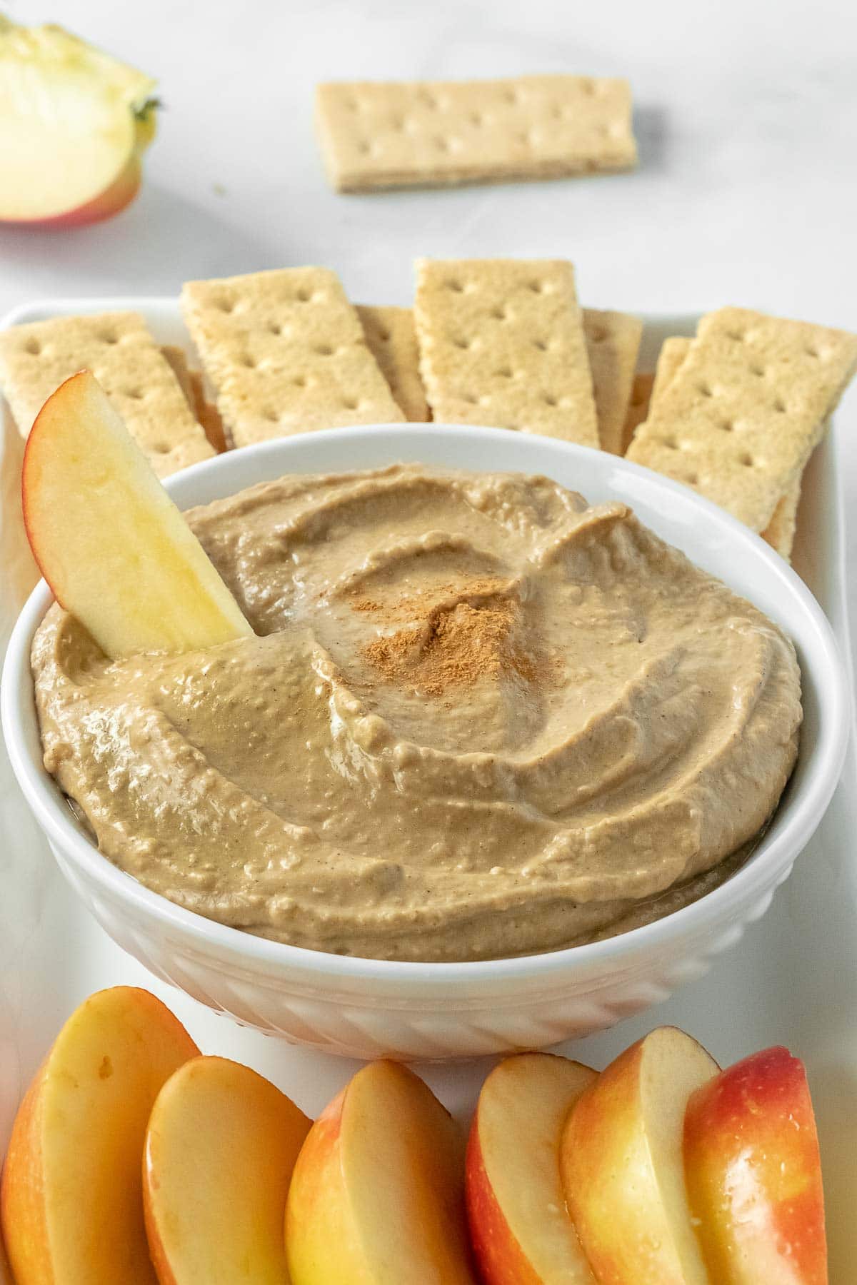 hummus in a shallow white bowl surrounded by apple slices and graham crackers