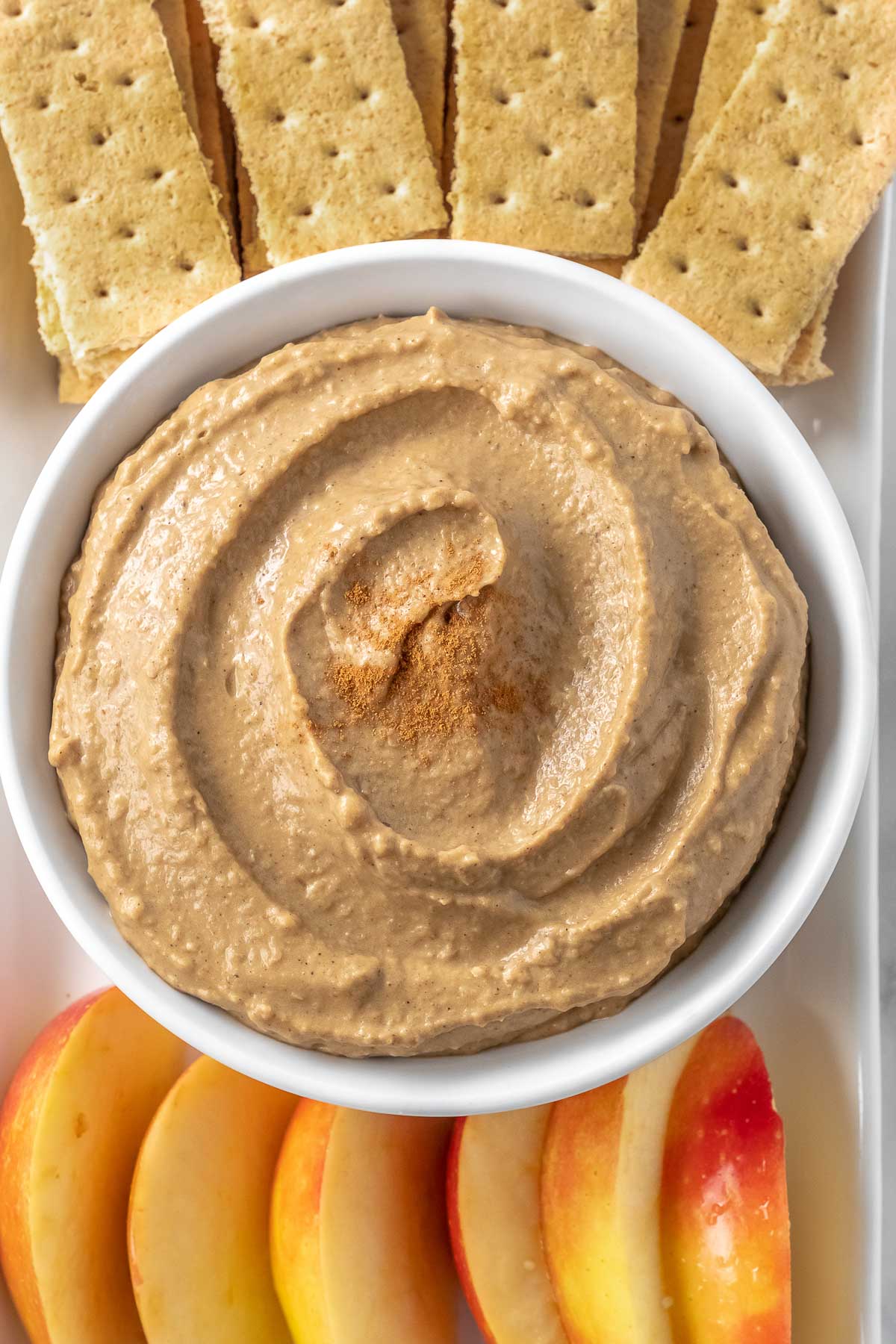closeup of hummus dip in a white bowl with apple slices and crackers on a plate