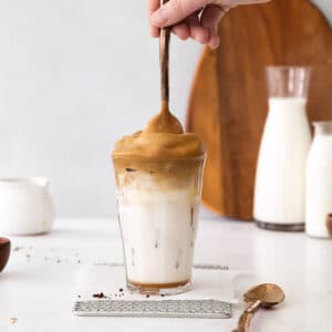 tall glass of whipped coffee with a spoon dipped in on a white counter with carafes of milk and wood cutting board, dalgona coffee