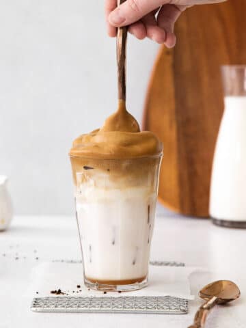 tall glass of whipped coffee with a spoon dipped in on a white counter with carafes of milk and wood cutting board, dalgona coffee