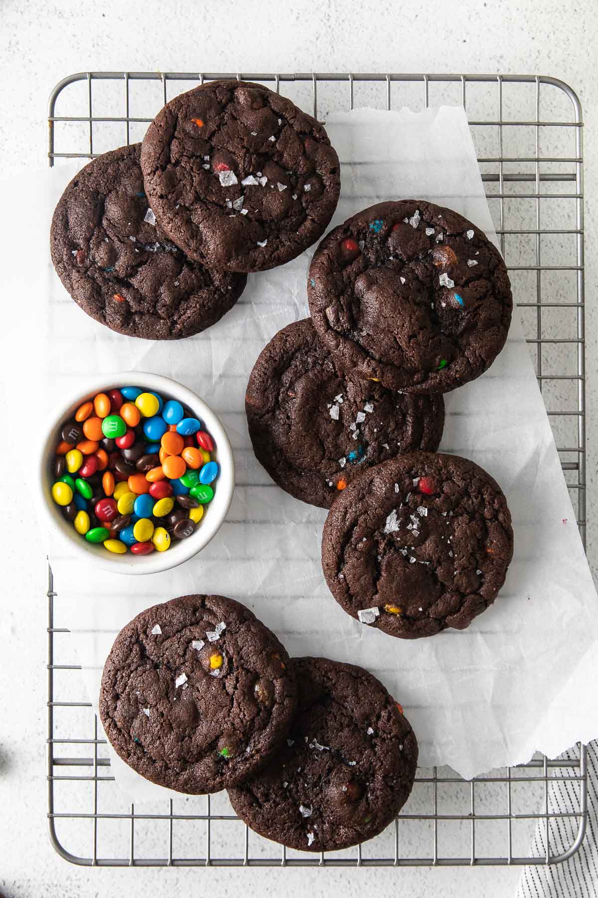 seven double chocolate chip cookies with m&m's on a wire rack with a small bowl of m&m's