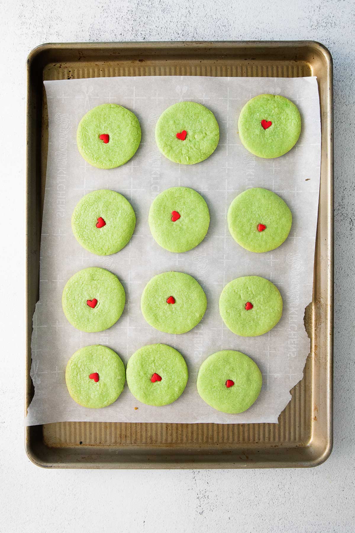 twelve green sugar cookies with a red heart in the center of each cookie on a parchment lined baking sheet