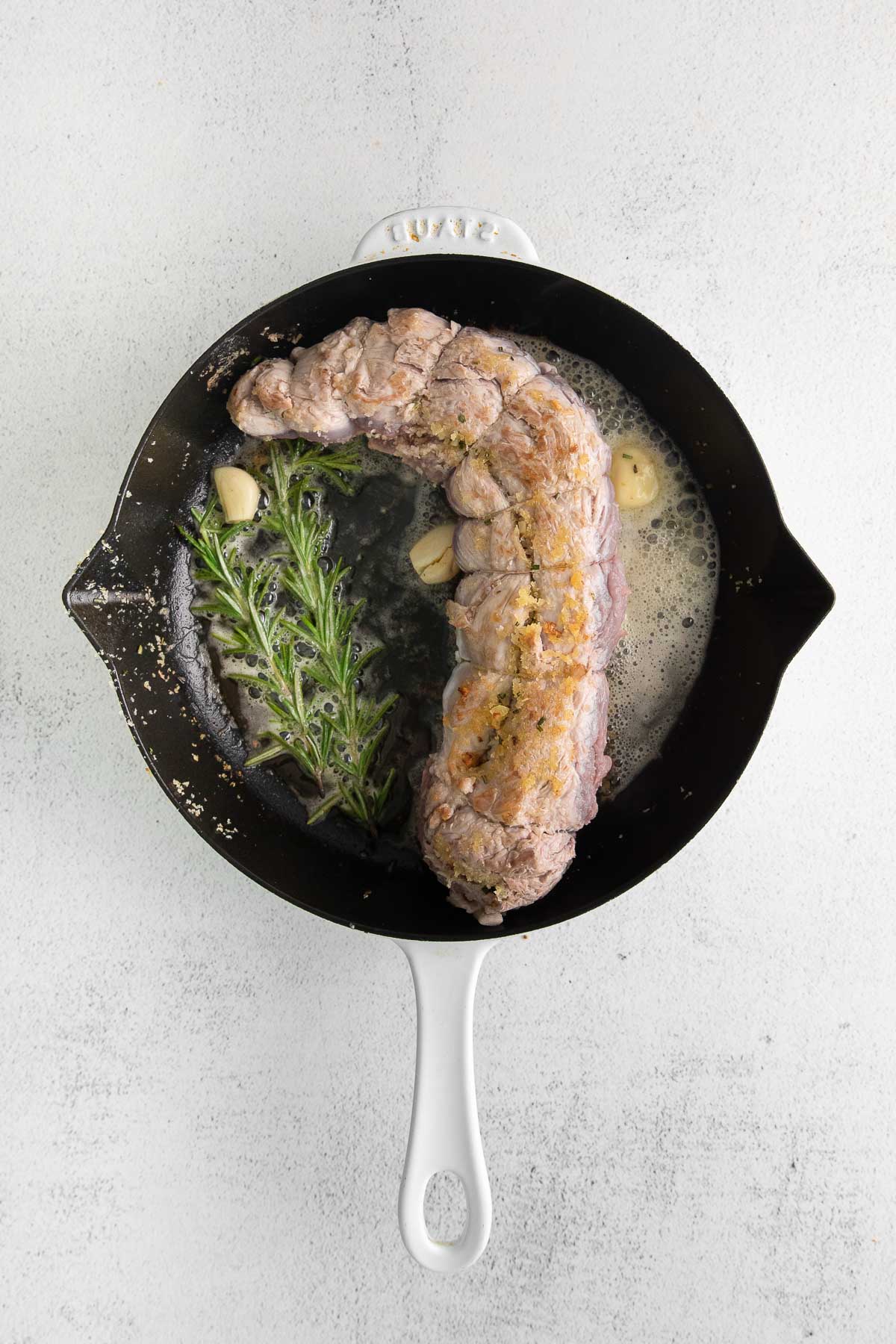pork tenderloin cooking in a cast iron pan with garlic cloves and rosemary sprigs