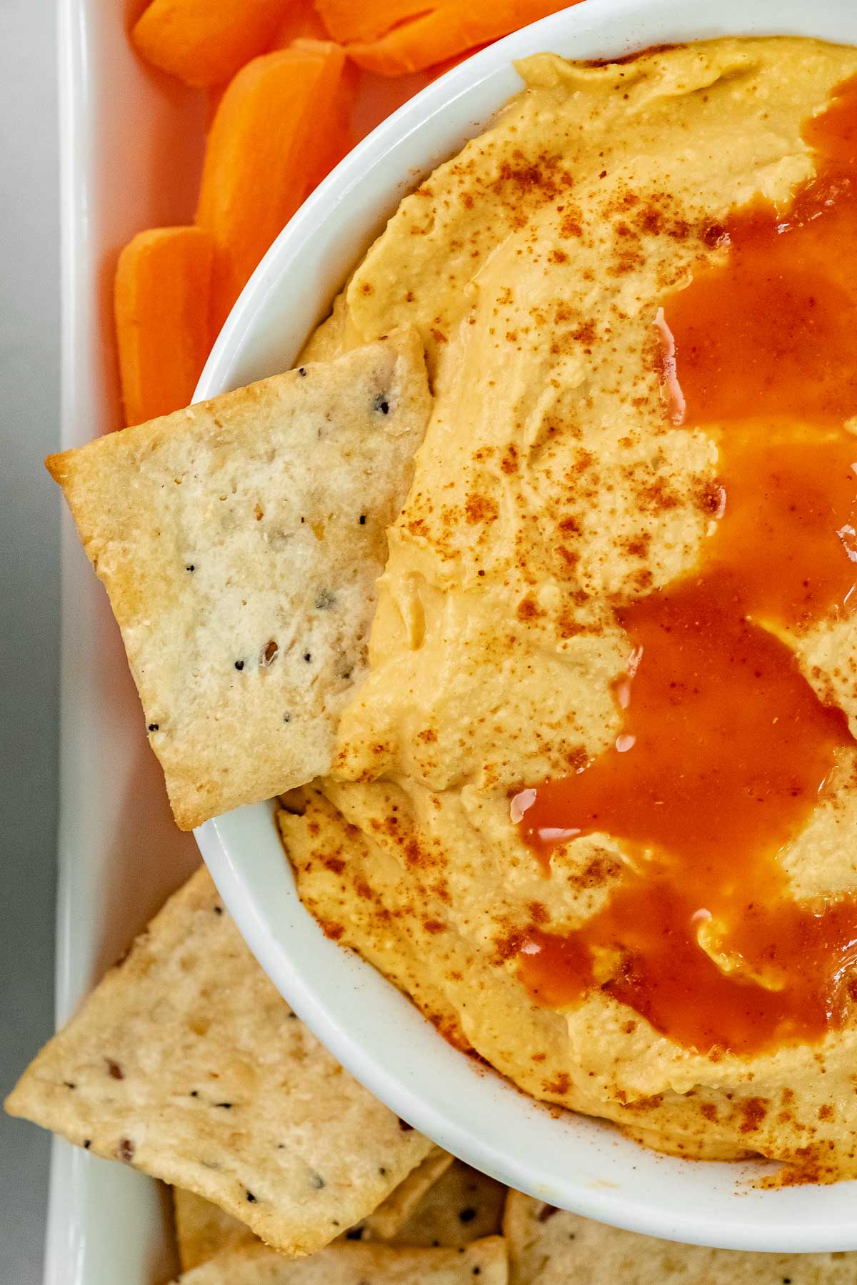 white bowl of hummus drizzled with buffalo sauce with a cracker dipped in it